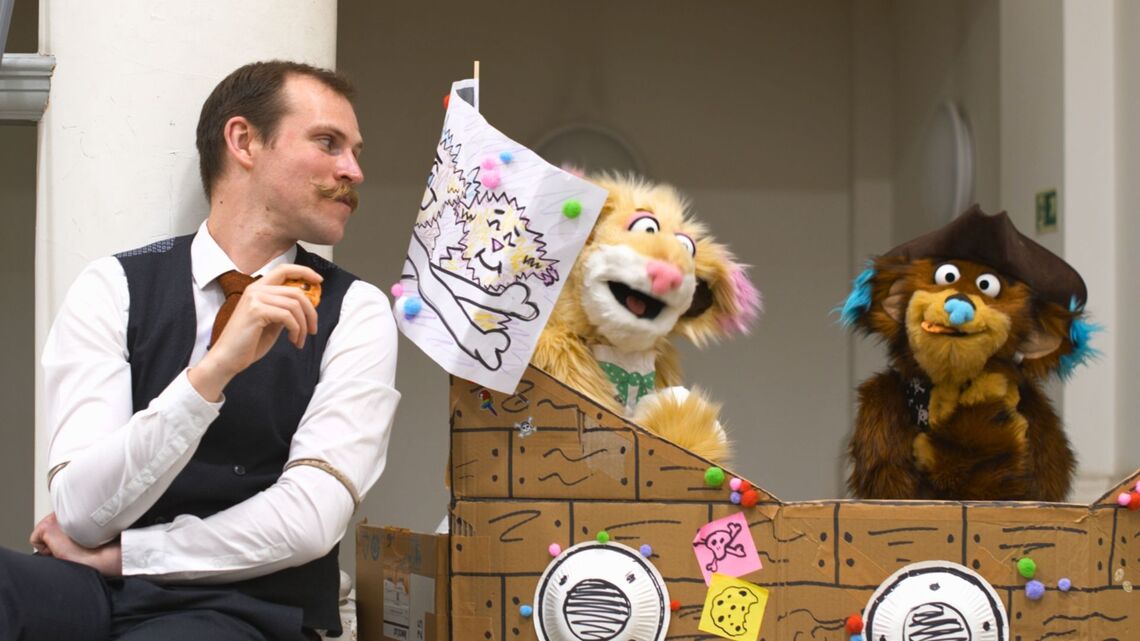 A photo of the overtheres - 2 puppet creatures - in a cardboard pirate ship. Their librarian friend is sat next to the boat, sharing cookies with them